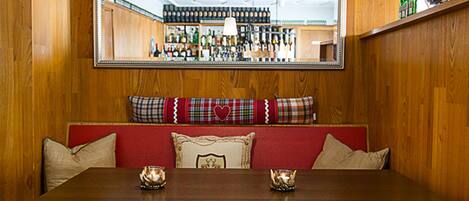 A wooden-paneled room with a red upholstered bench, two candles, and a mirror reflecting a bar area.