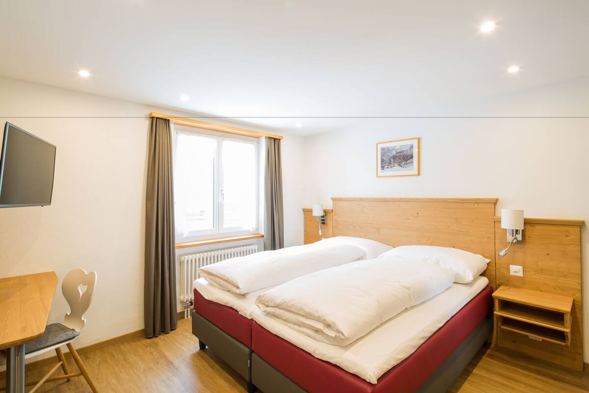 A hotel room with a bed, a desk, a chair, a window with curtains, a radiator, and a picture on the wall.