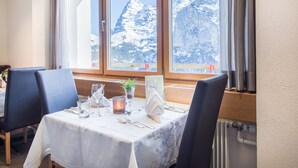 A dining table set with a candle, glassware, and a menu stand, with a mountain view through large windows. - Hotel Edelweiss Superior (Mürren)