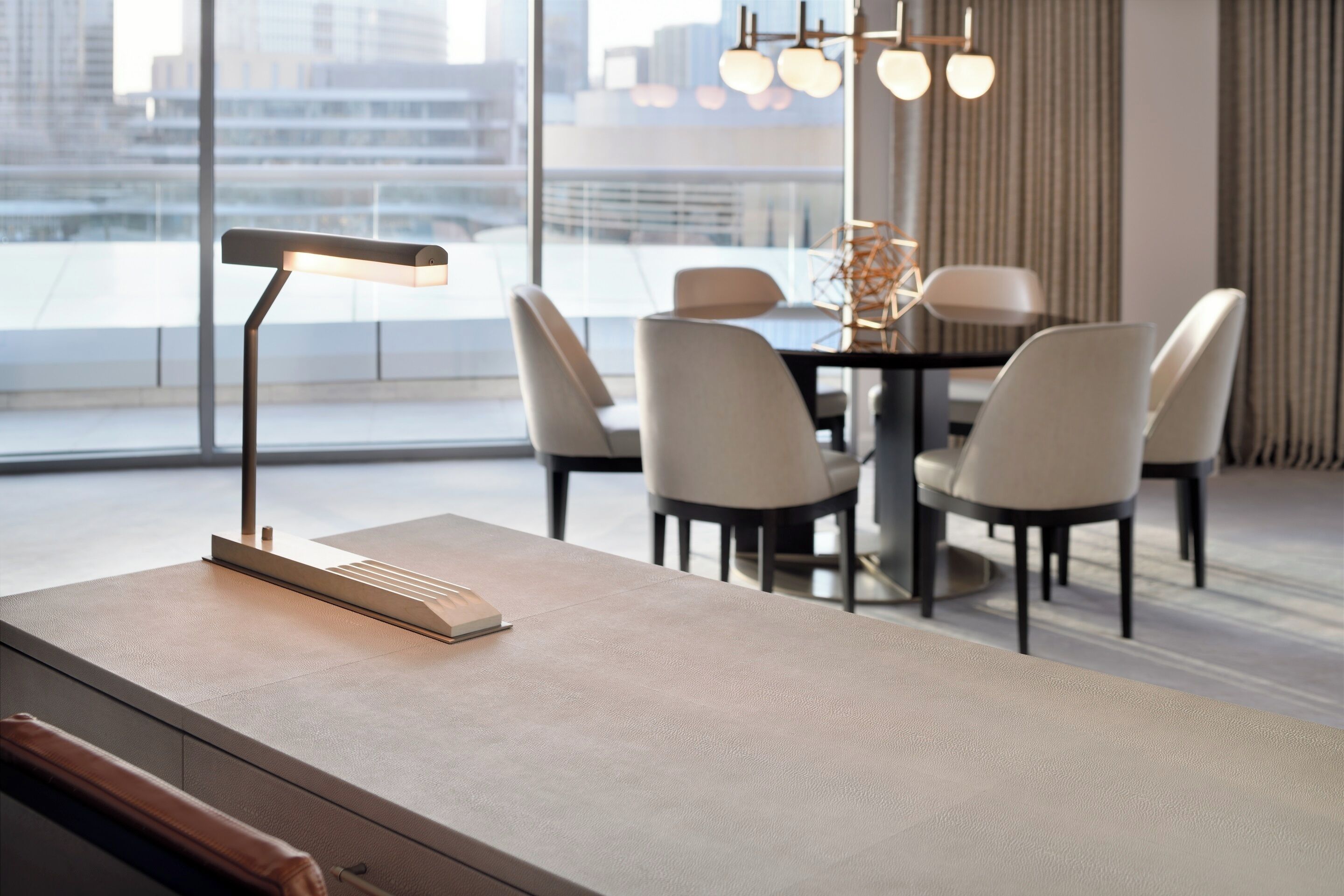 Un área de comedor moderna con una mesa y sillas minimalistas, una lámpara de pie y vistas a la ciudad a través de amplias ventanas.