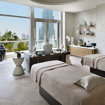 A spa room with two massage tables, a large window with a city view, and a decorative shelf with various items.