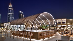 A modern bar area with a curved roof structure, seating arrangements, and a view of the city skyline at night.