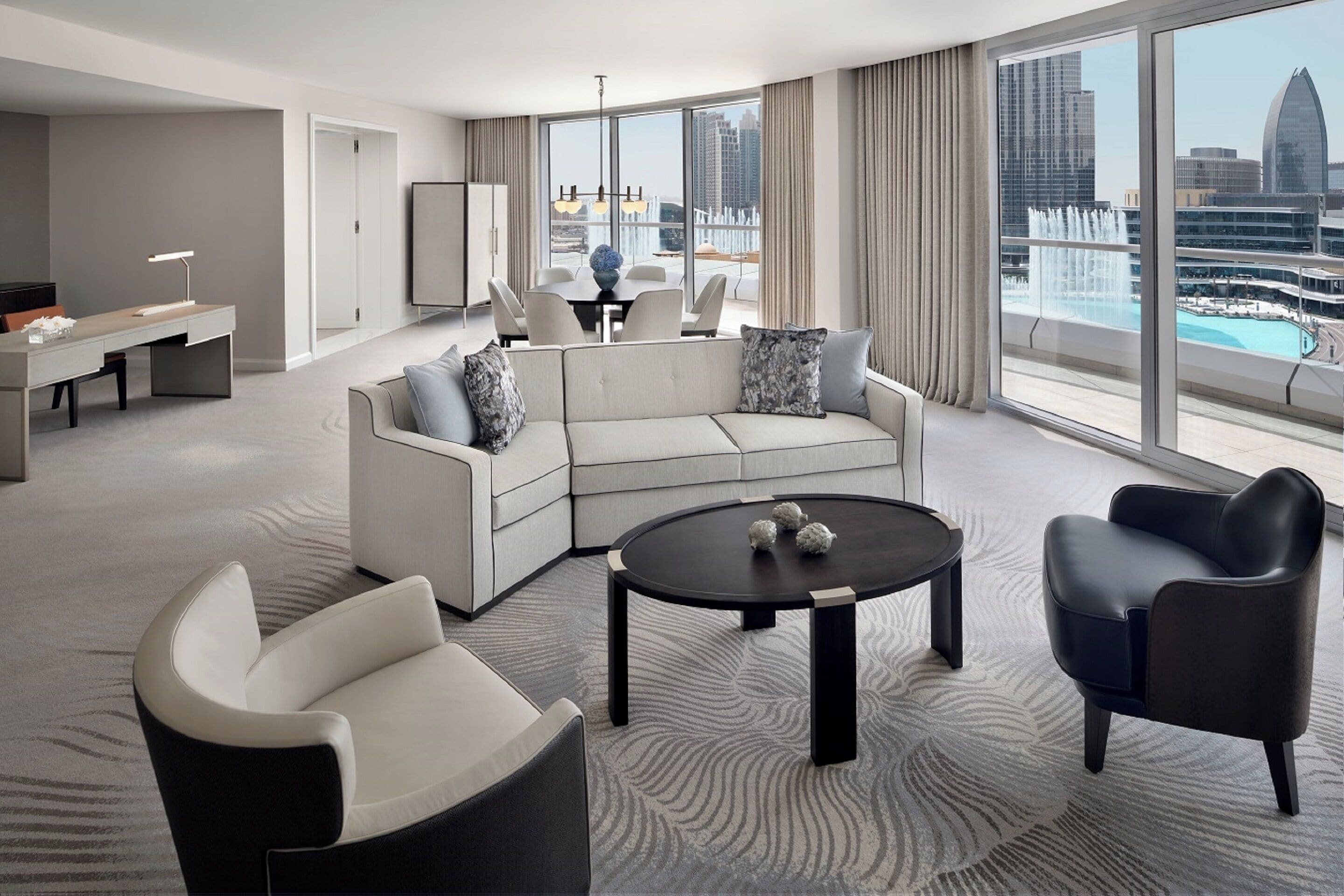 A modern hotel room with a large window, a sofa, a coffee table, and a desk with a lamp.