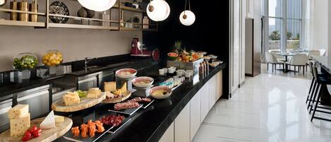 A modern kitchen with a black countertop displaying various food items, a coffee machine, and a view of the dining area through large windows.