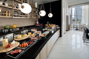 A modern kitchen with a black countertop displaying various food items, a coffee machine, and a view of the dining area through large windows.