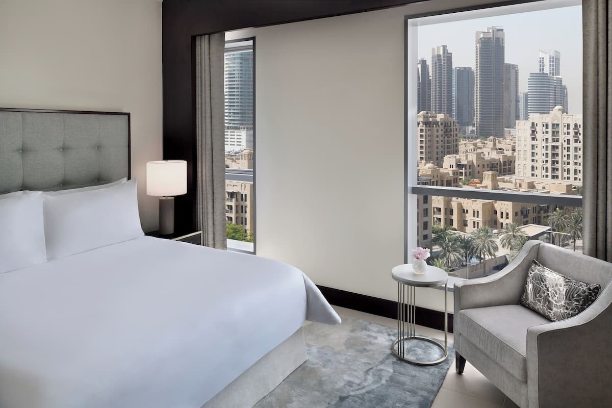 A hotel room with a bed, a chair, a small table with a lamp, and a view of a cityscape through large windows.