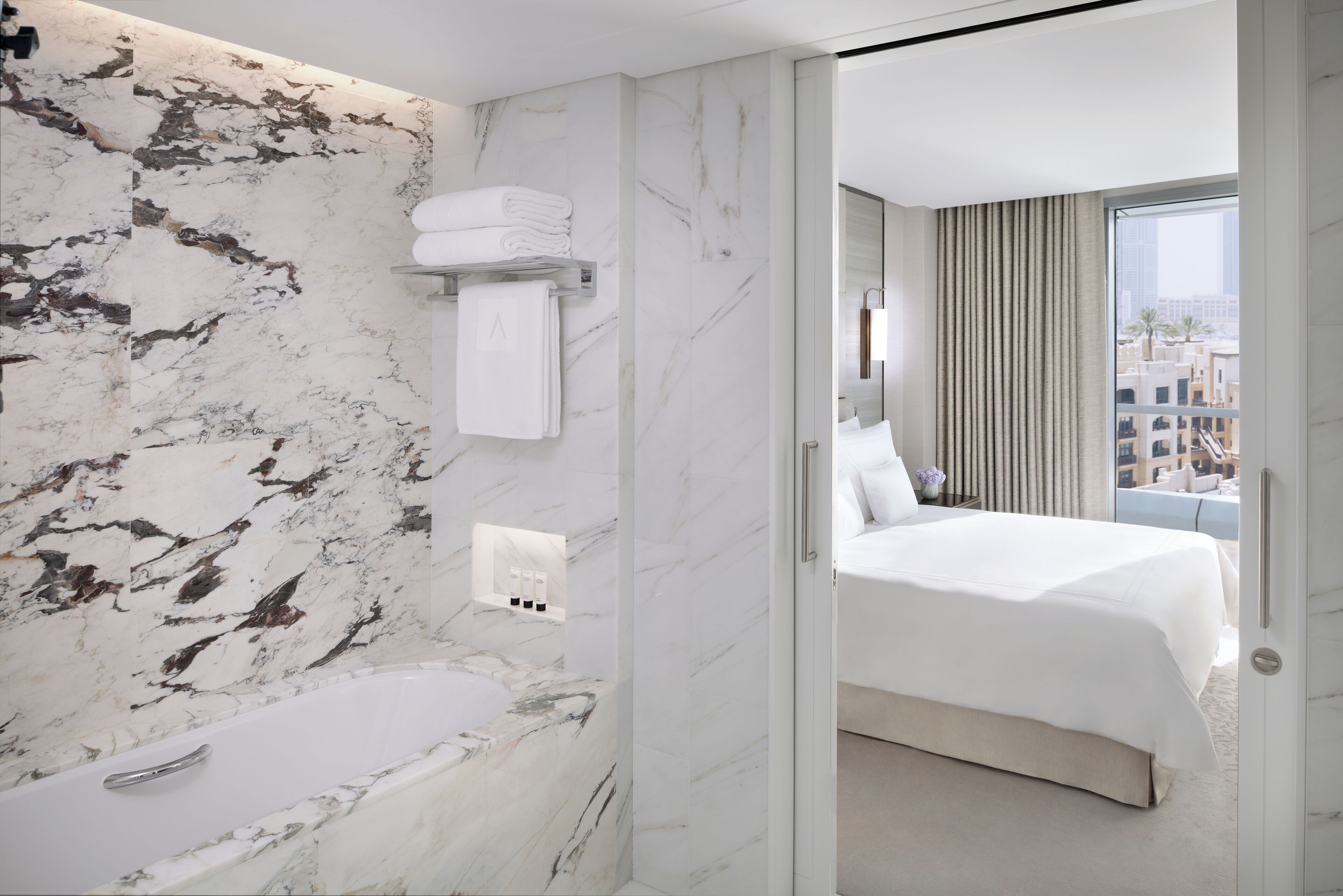 A modern hotel room with a marble bathroom, a bed with white linens, and a view of the city through large windows.
