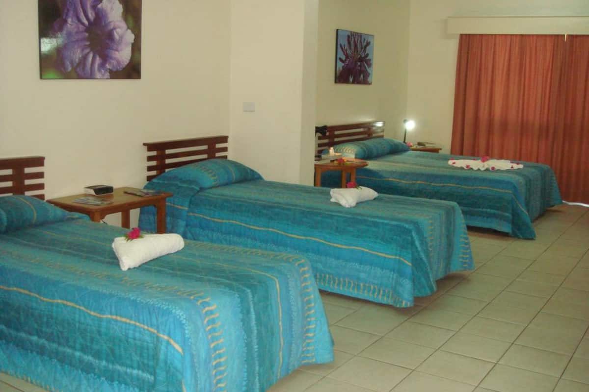 A hotel room with three beds, each with a blue bedspread and a white towel on one of them. There are small tables with lamps and a painting on the wall.
