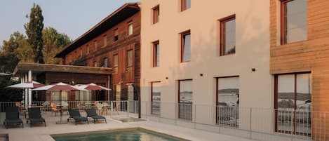 Un área de piscina con sillones de descanso y sombrillas, un edificio con amplias ventanas y vistas al paisaje.