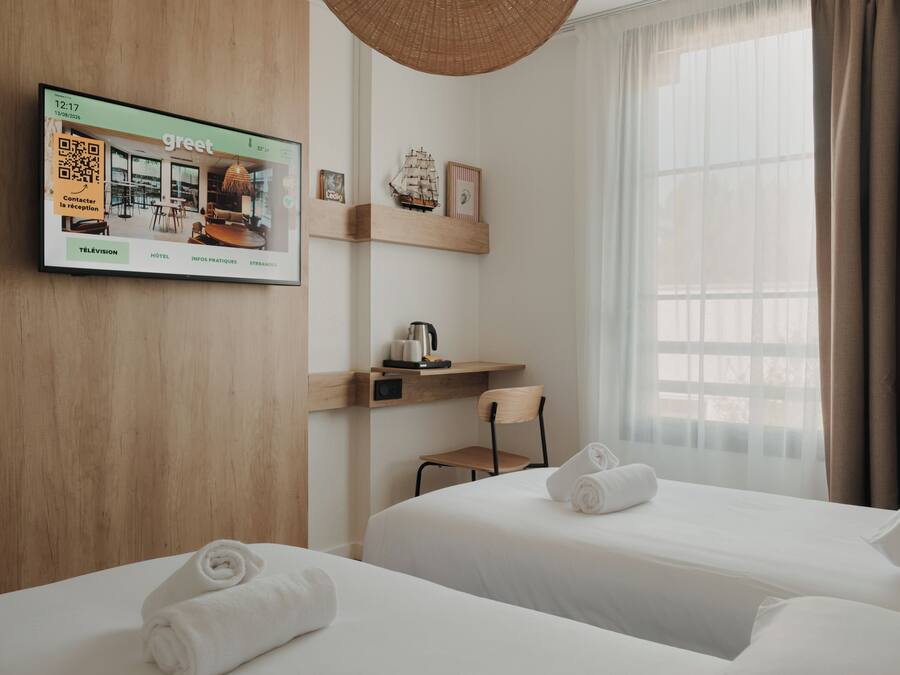 Quarto de hotel com duas camas, televisão na parede, mesa com chaleira e janela com cortinas.