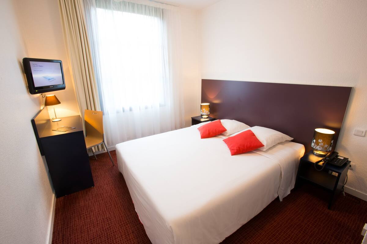 A hotel room with a bed, bedside lamps, a TV, and a window with curtains.