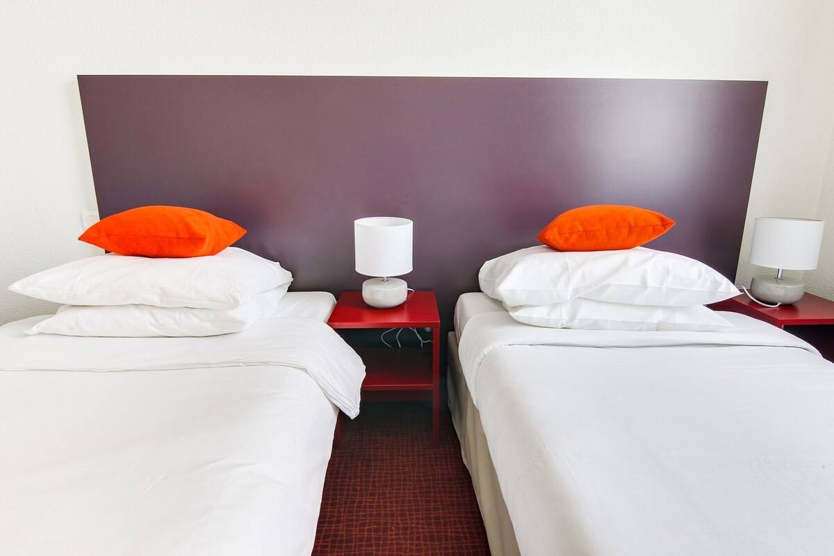 A hotel room with two beds, each with white bedding and orange pillows, flanked by red nightstands with white lamps.