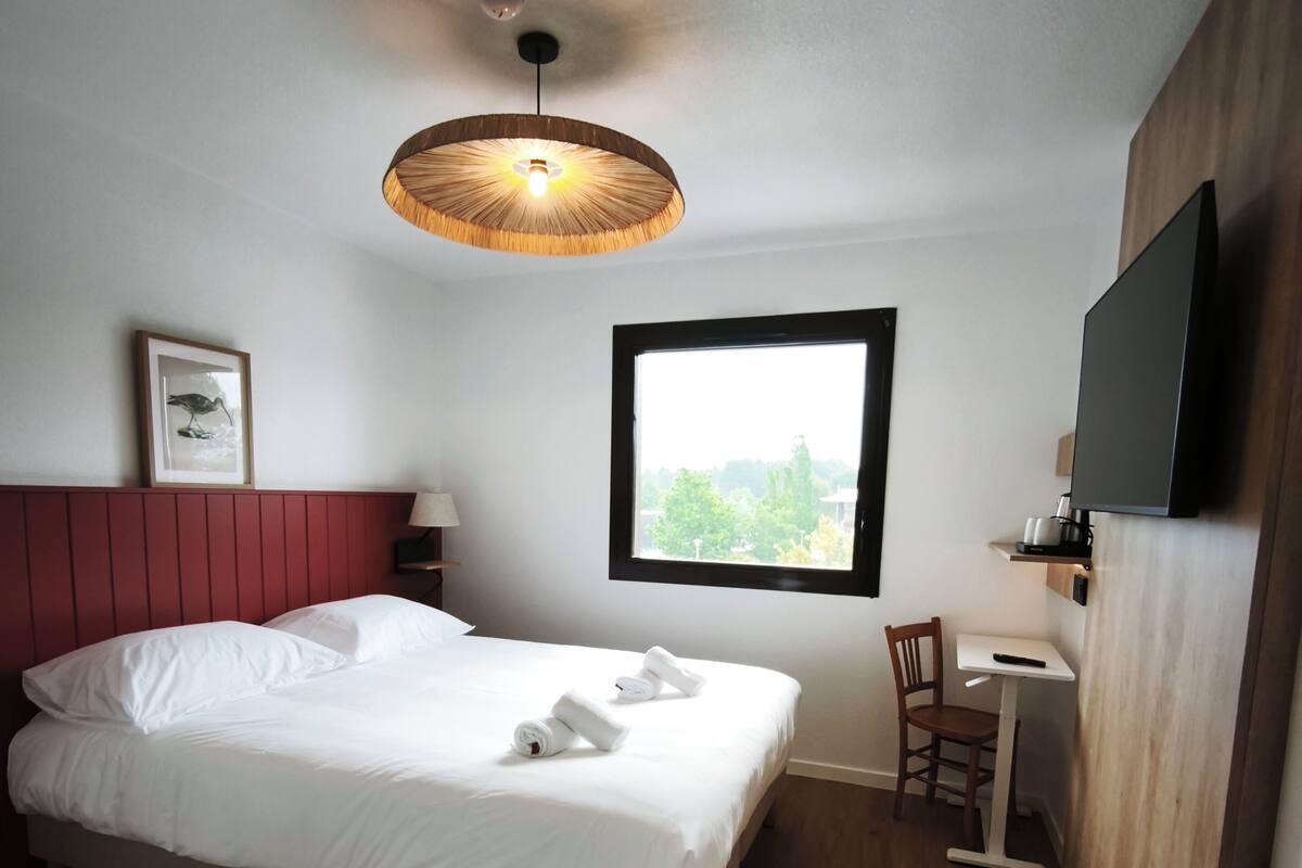 A bedroom with a bed, a window, a desk, a chair, a TV, and a ceiling light.