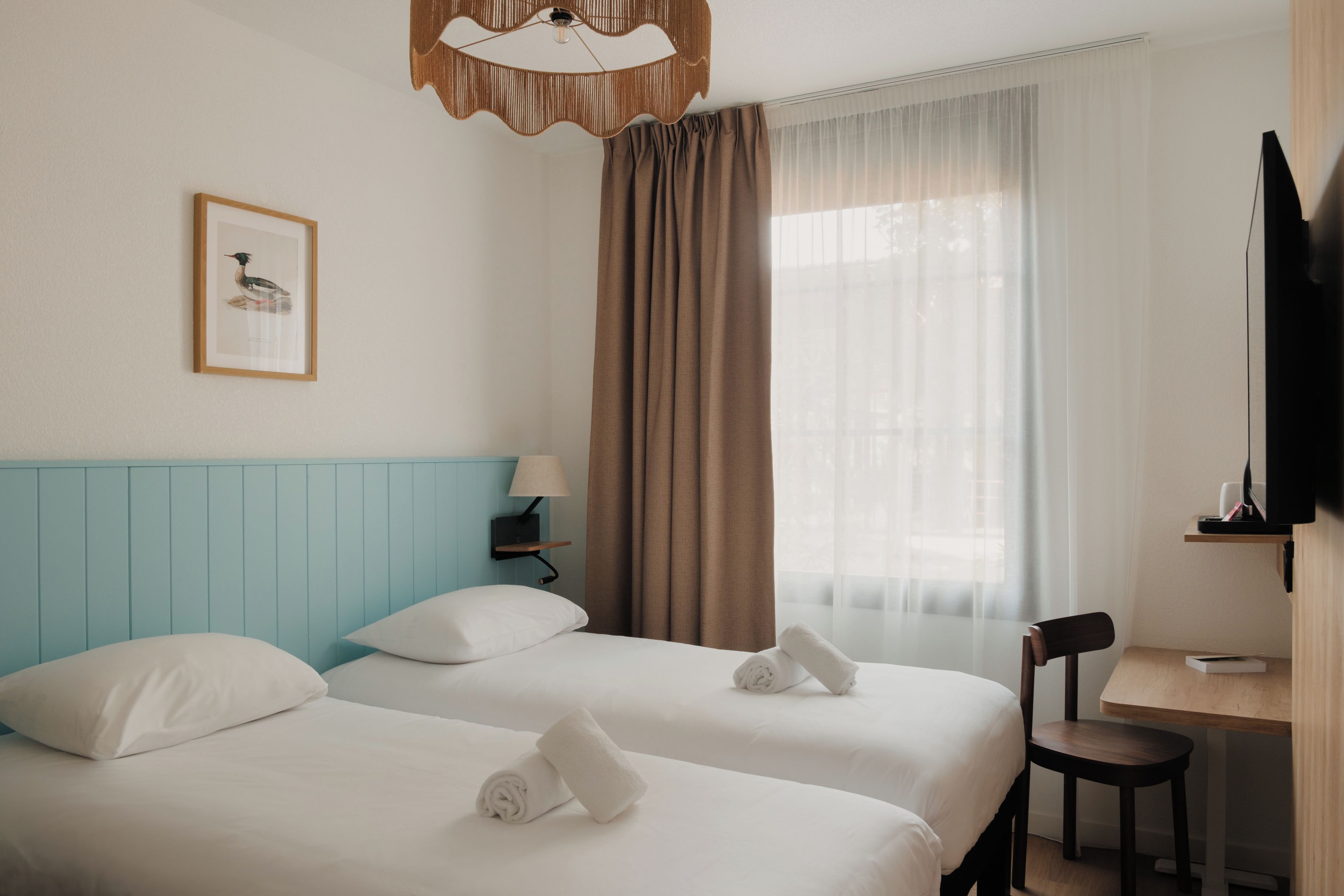 Une chambre d’hôtel avec deux lits, un bureau, une chaise, une télévision, une lampe, un cadre photo et une fenêtre avec des rideaux.