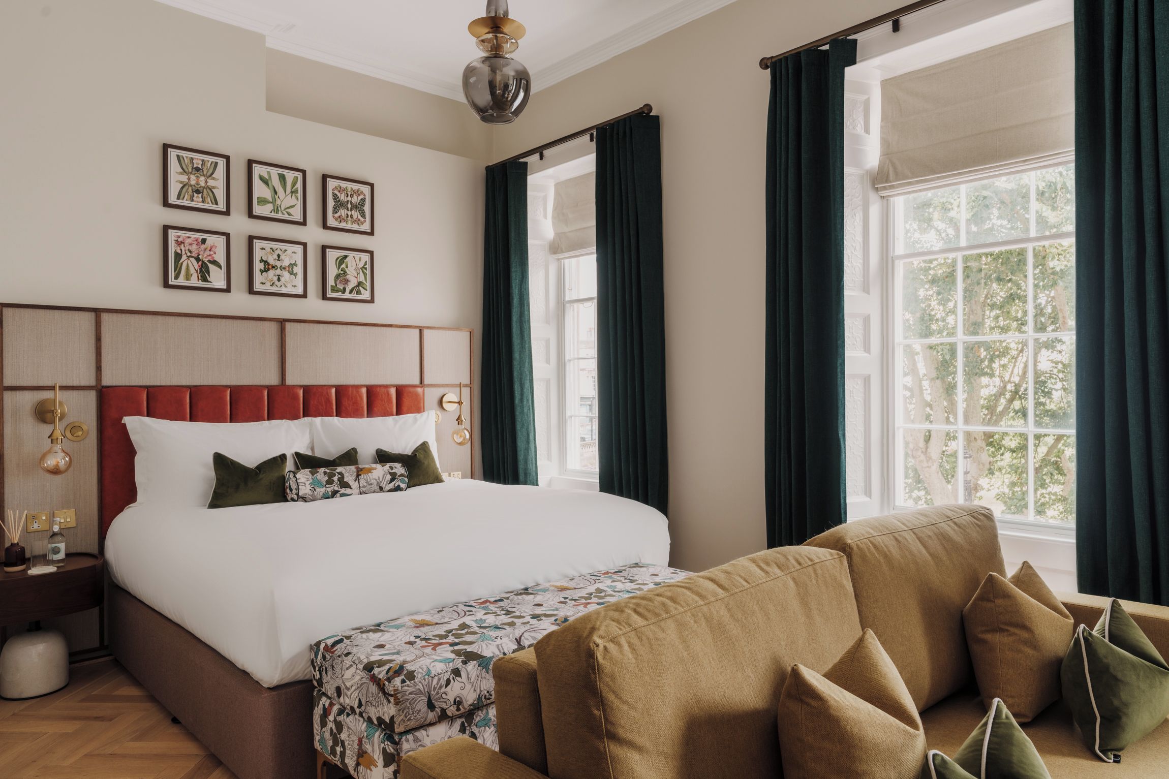 Quarto de hotel com uma cama grande, um sofá, uma mesinha de cabeceira, uma luminária e uma janela com cortinas.