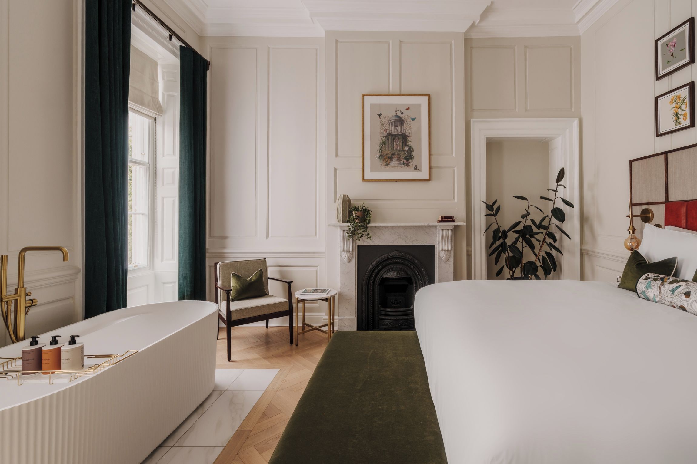 A hotel room with a large bed, a sitting area with a chair, a fireplace, and a bathtub with toiletries.