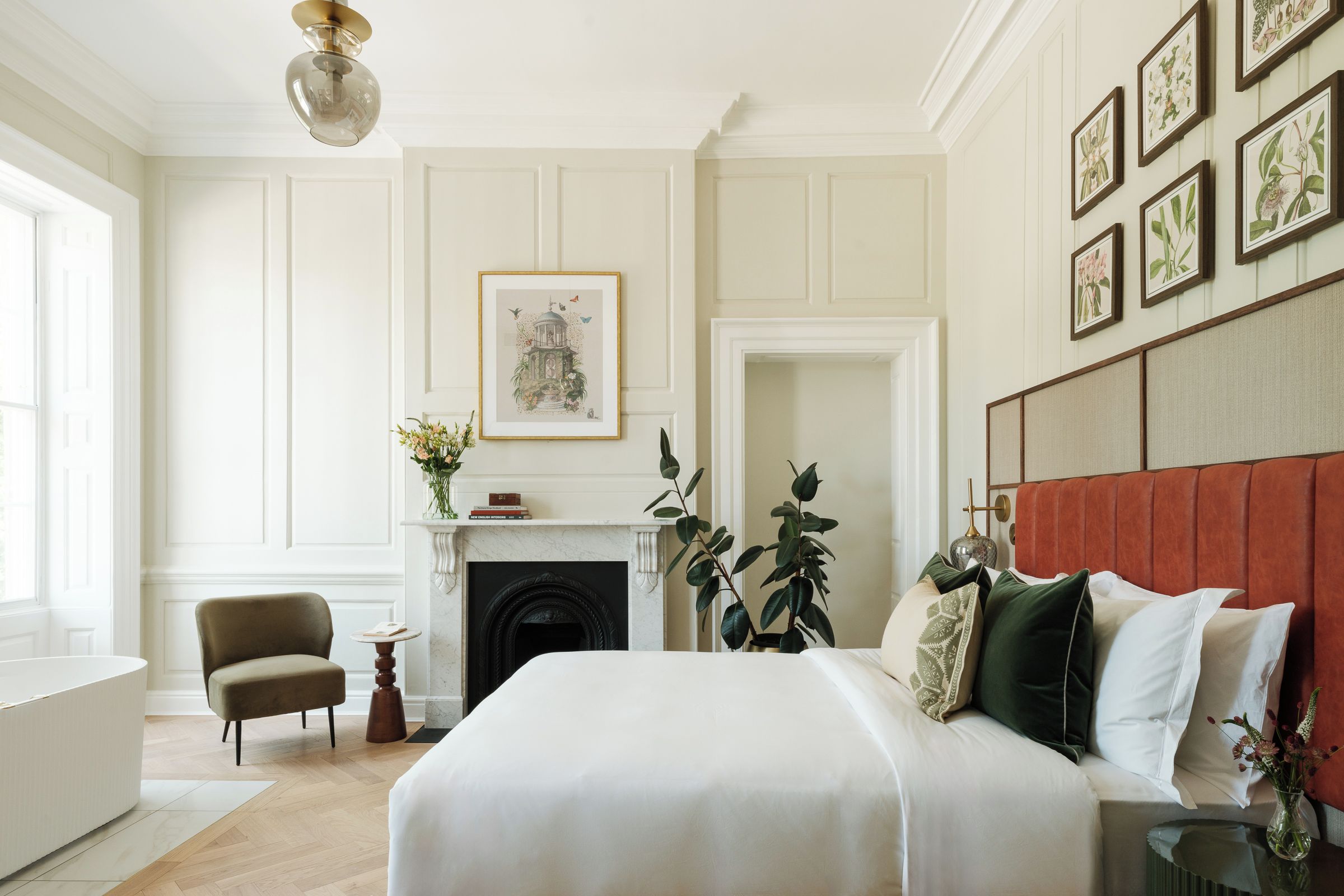 A bedroom with a large bed, a fireplace, a chair, and a wall with framed botanical prints.