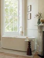 A bathroom with a freestanding bathtub, a round table with a glass of amber liquid, and a vase with flowers on a mantelpiece.