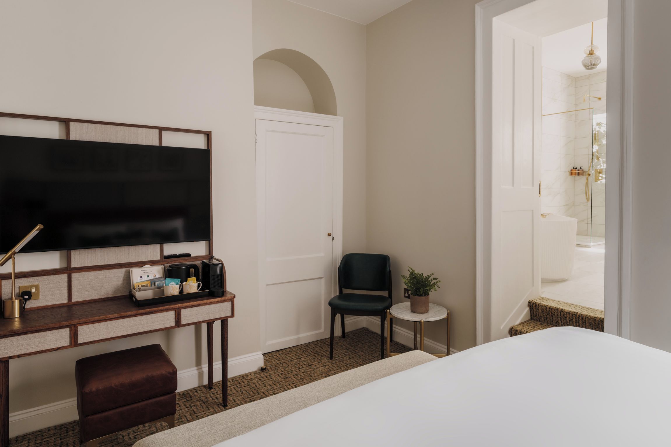A hotel room with a bed, a desk with a flat-screen TV, a chair, a small table with a plant, and a view of the bathroom.