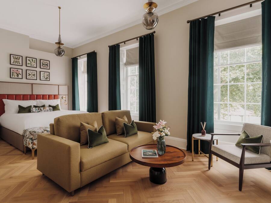 Quarto de hotel com cama, sofá, poltrona, mesa de centro redonda e amplas janelas com cortinas.