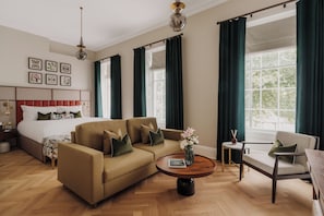 Quarto de hotel com cama, sofá, poltrona, mesa de centro redonda e amplas janelas com cortinas.