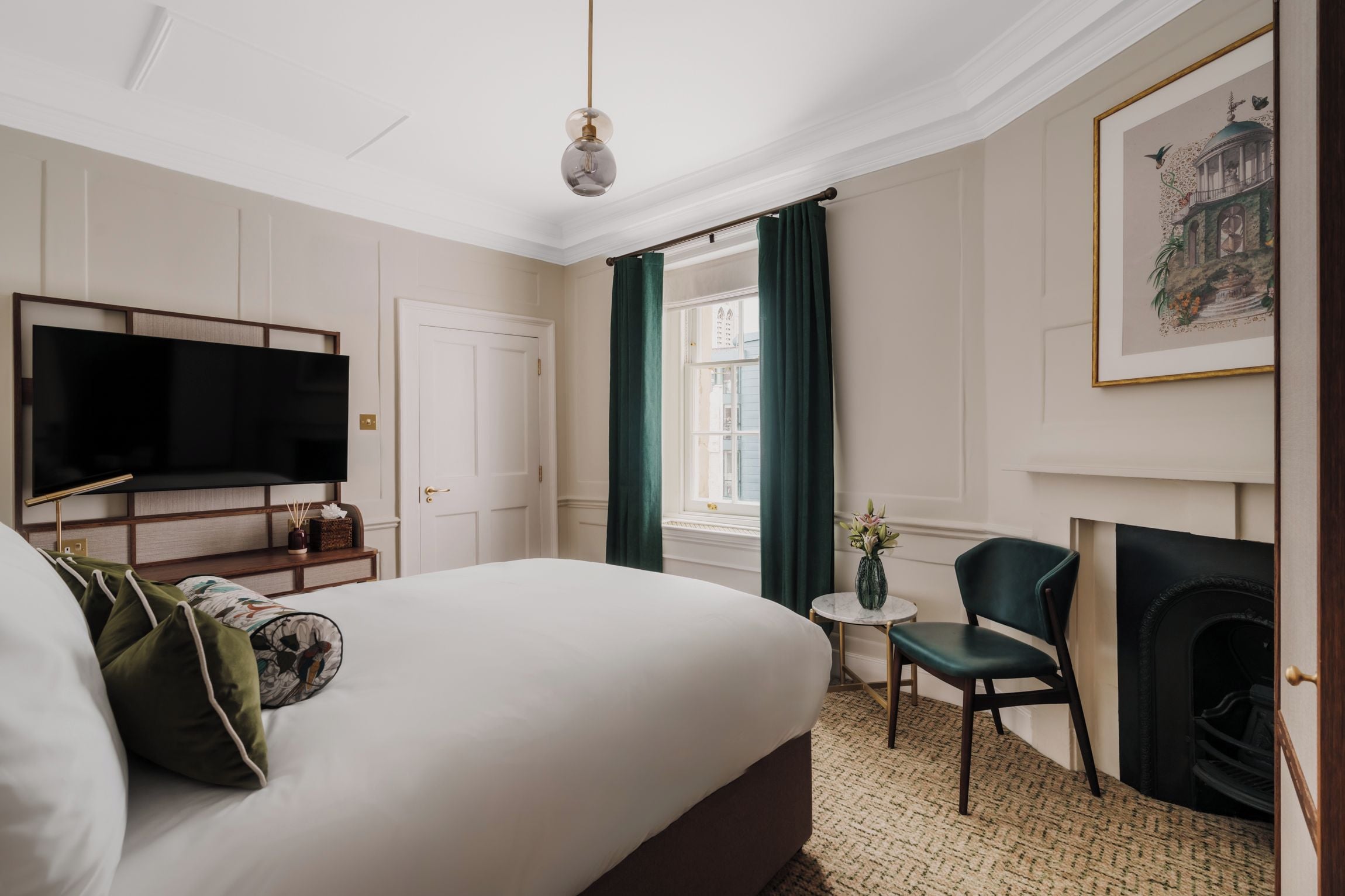 A hotel room with a large bed, a television, a small table with a vase, a chair, and a fireplace.