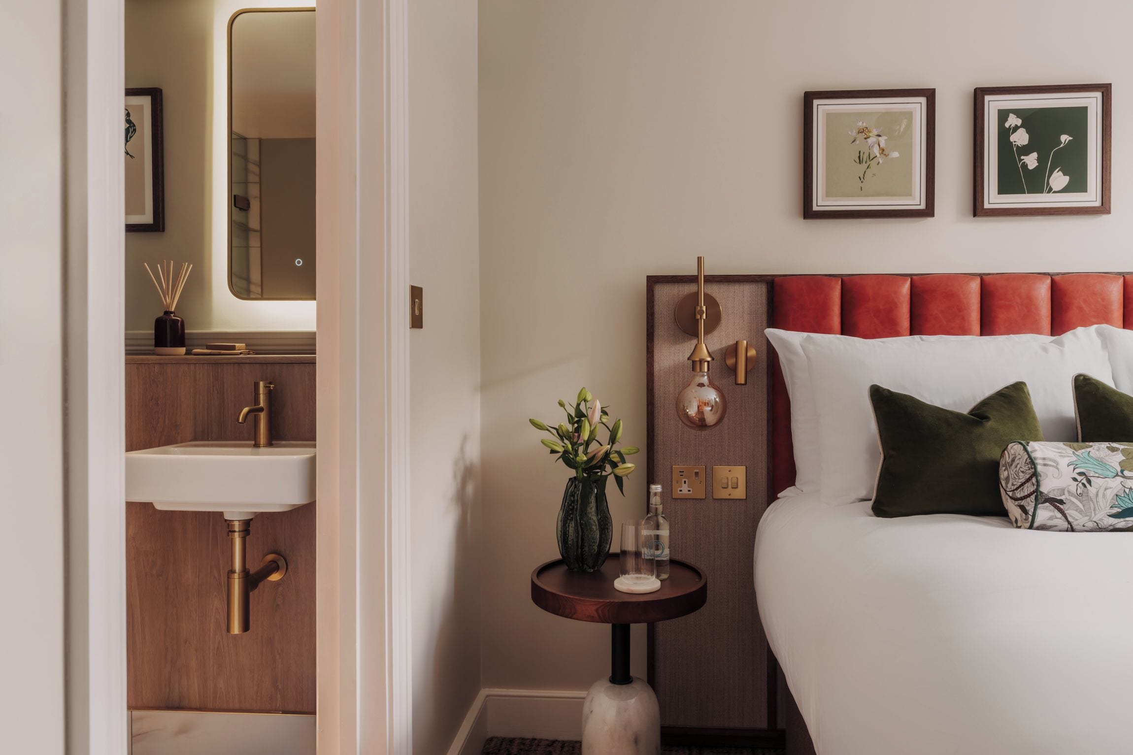 Ein Hotelzimmer mit einem Bett, einem Nachttisch mit Blumenvase, einer Wanduhr und einem Spiegel.