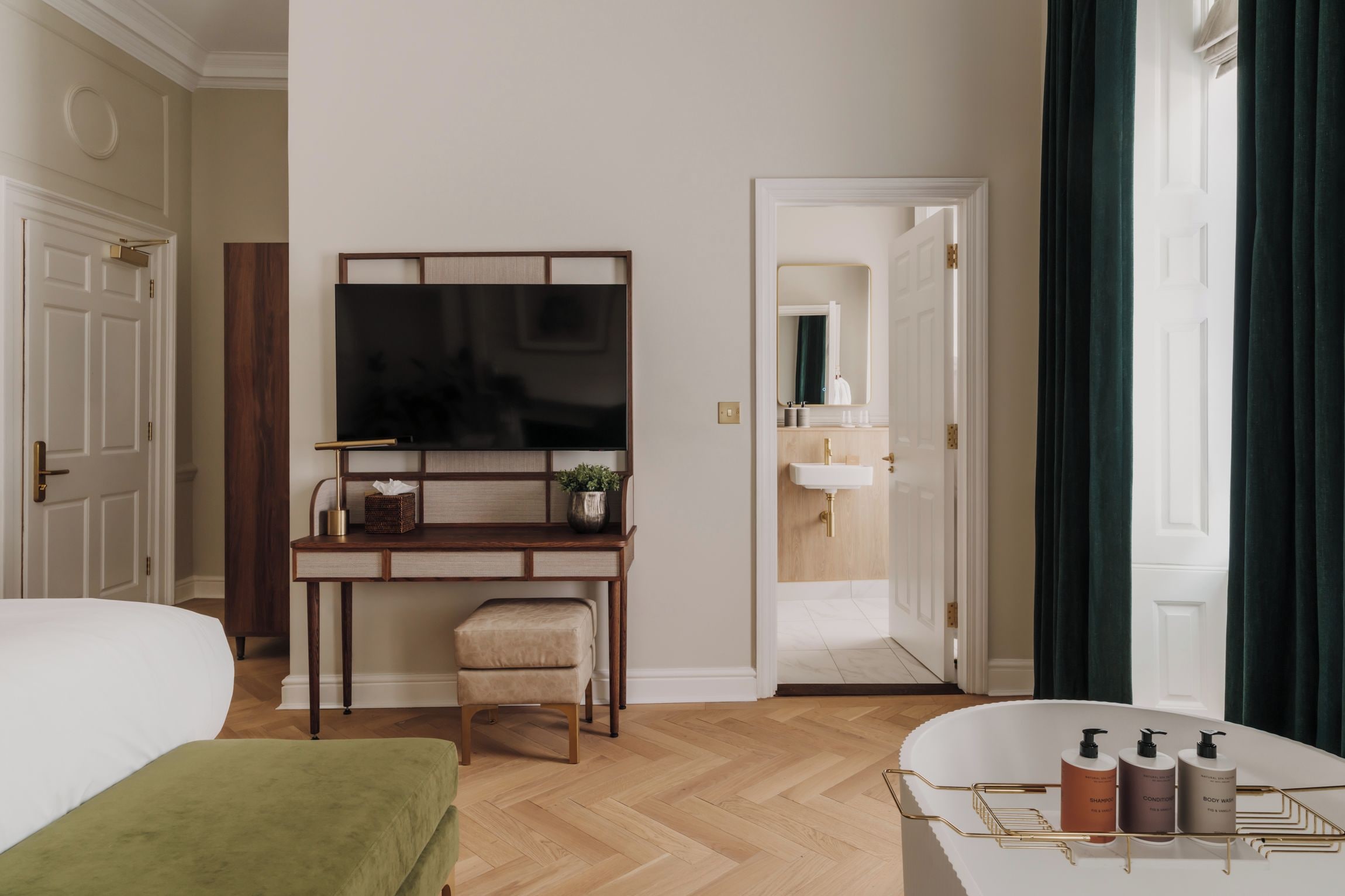A hotel room with a bed, a desk with a flat-screen TV, a chair, a small stool, a vanity with a mirror, and a bathtub with toiletries.