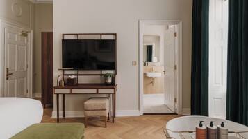 A hotel room with a bed, a desk with a flat-screen TV, a chair, a small stool, a vanity with a mirror, and a bathtub with toiletries.