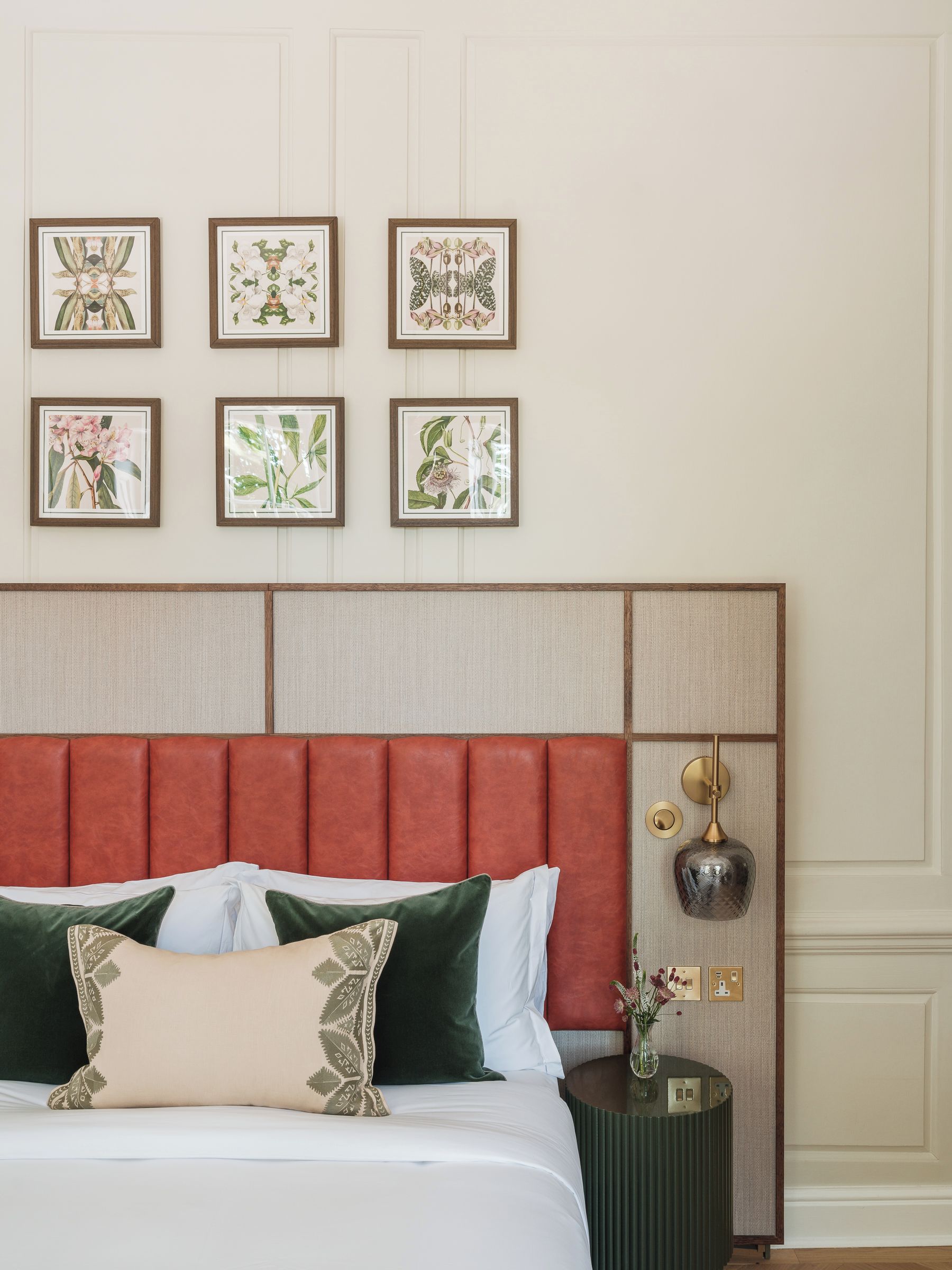 A hotel room with a bed, a nightstand, a vase with flowers, and six framed botanical prints on the wall.