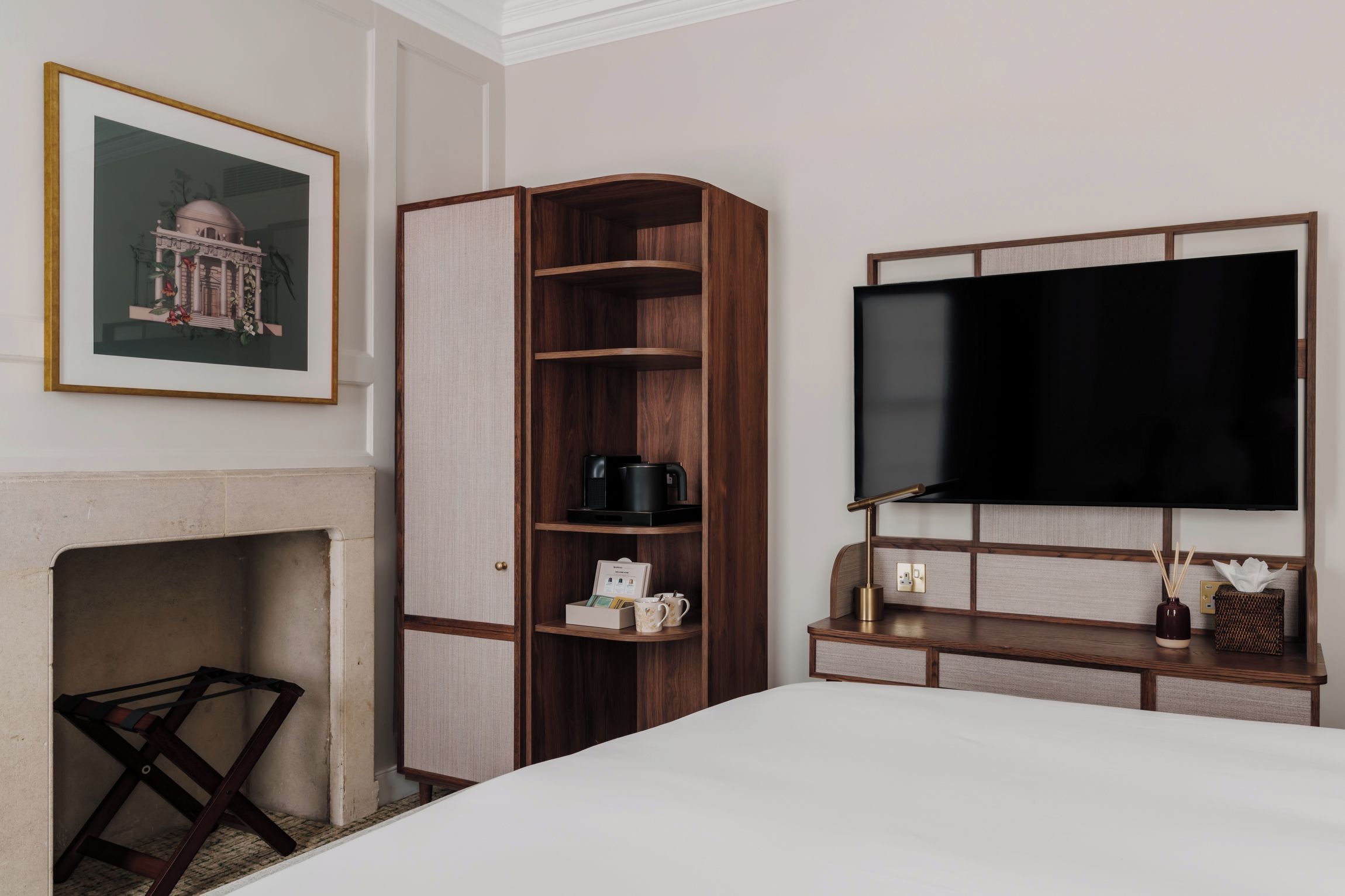 Habitación de hotel con una cama, un televisor montado en la pared, un mueble de madera con estantes, una chimenea y un cuadro enmarcado.