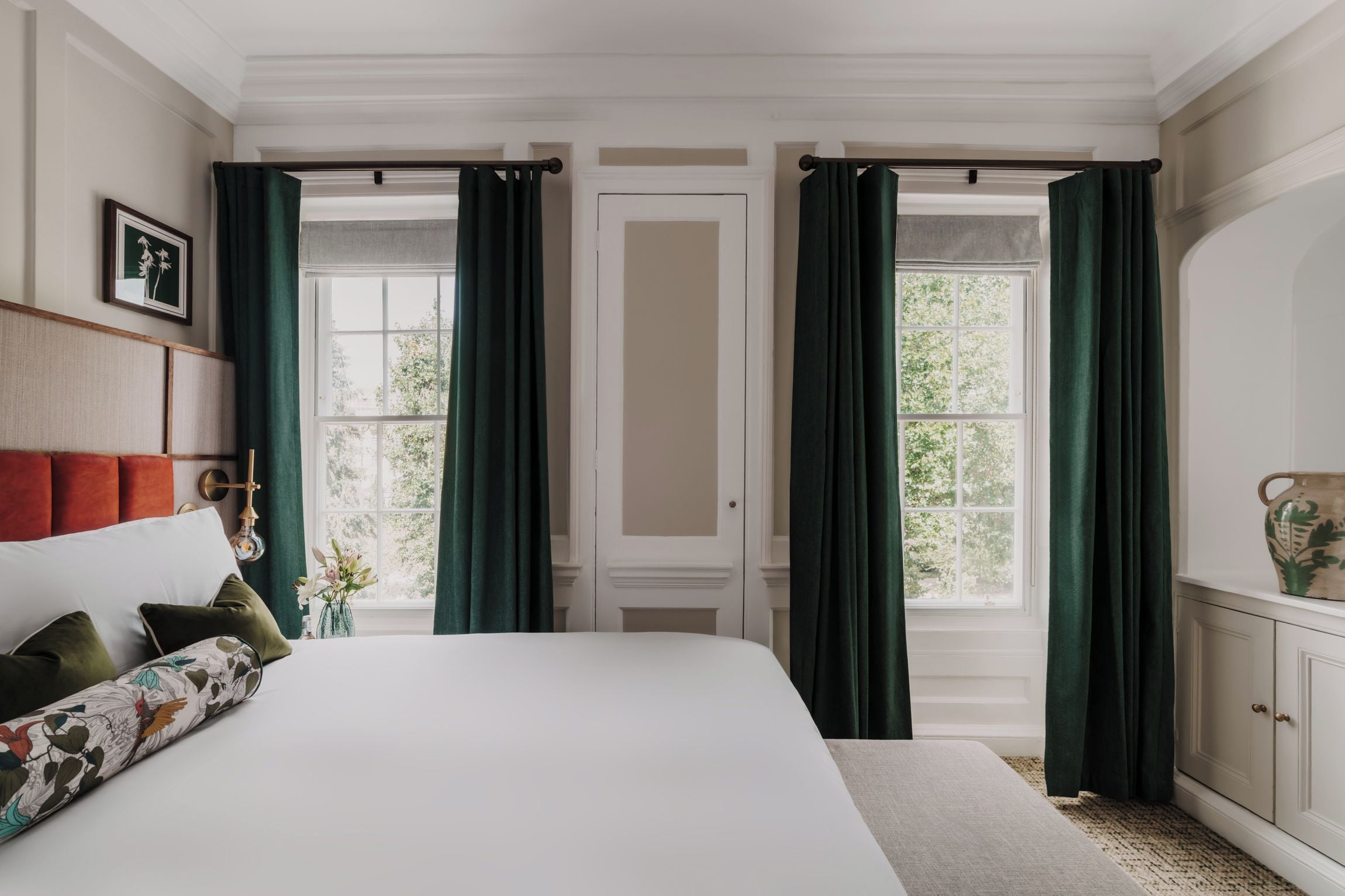 Un dormitorio con una cama grande, una mesita de noche, un tocador y dos ventanas con cortinas.