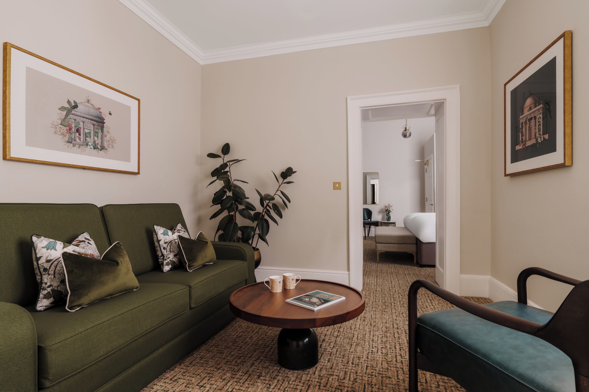 Uma sala de estar com um sofá verde, uma mesa de centro e duas poltronas. Há quadros nas paredes, uma planta e uma porta que dá para outro cômodo.