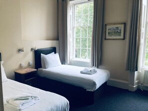 A hotel room with two beds, a window with curtains, a framed picture on the wall, and a small bedside table. - Parade Hotel Bath (Bath)