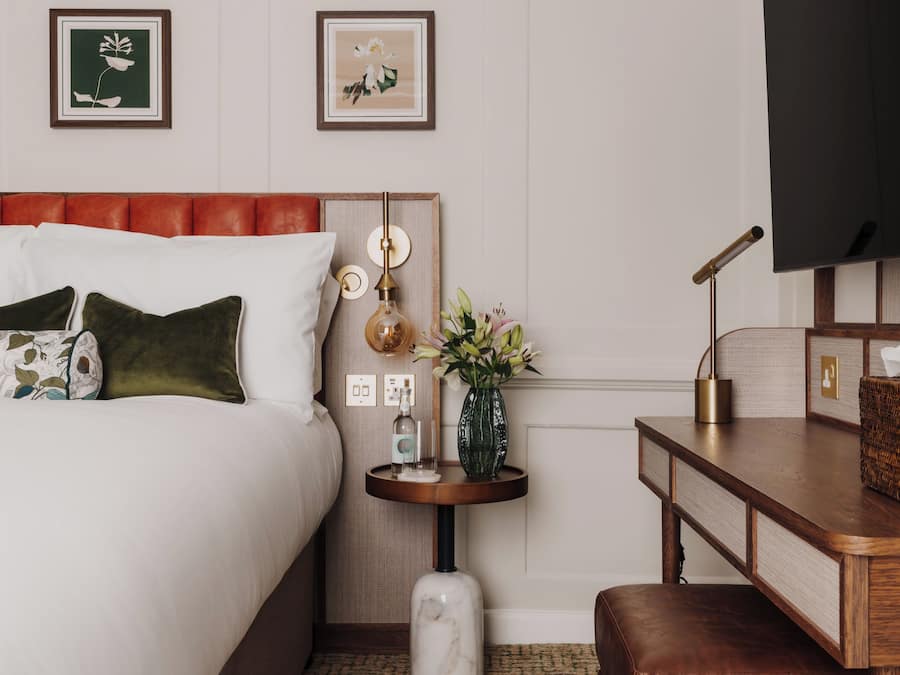 A hotel room with a bed, a bedside table with a vase of flowers, a lamp, and a television on the wall.