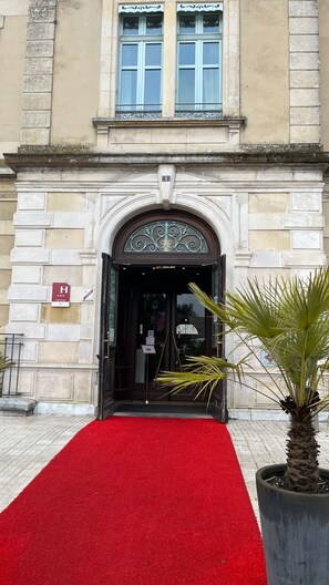 Entrada de un edificio con alfombra roja que conduce a una puerta oscura, una palmera a la derecha y un cartel con una clasificación por estrellas a la izquierda.