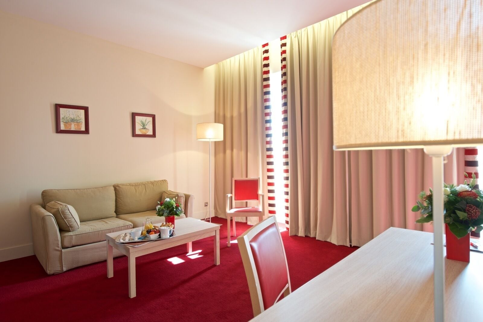 Habitació d'hotel amb una catifa vermella, un sofà beix, una taula petita amb una làmpada, una cadira vermella i una finestra amb cortines a ratlles.
