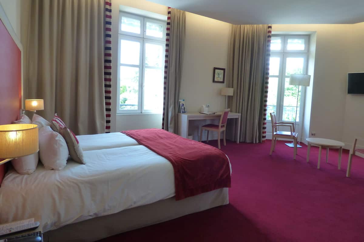 Quarto de hotel com cama, escrivaninha, cadeira, televisão e grandes janelas.