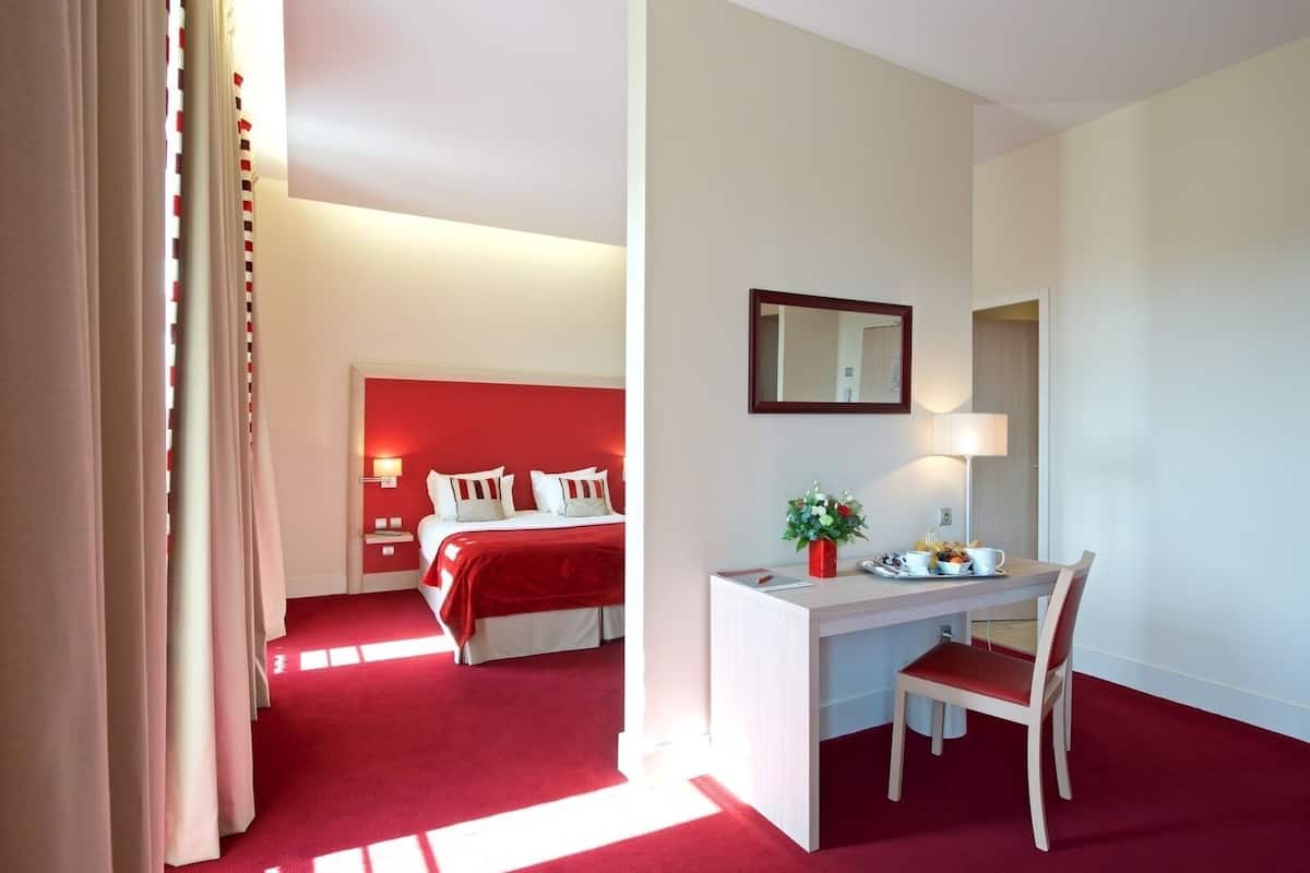Quarto de hotel com cama, escrivaninha e cadeira, espelho e mesa de cabeceira com abajur e flores.