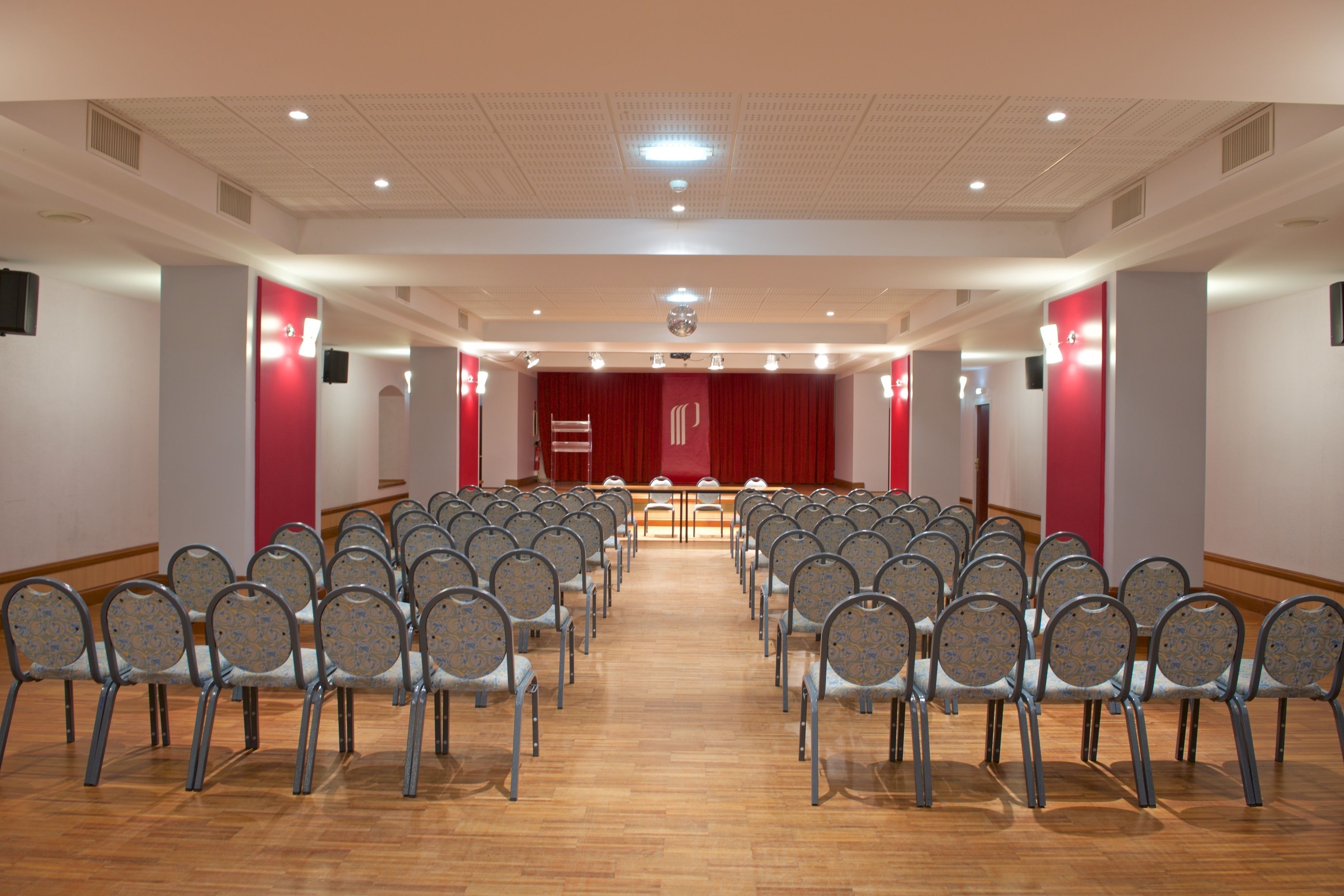 A conference room with rows of chairs facing a stage, wooden floors, and a red curtain backdrop.