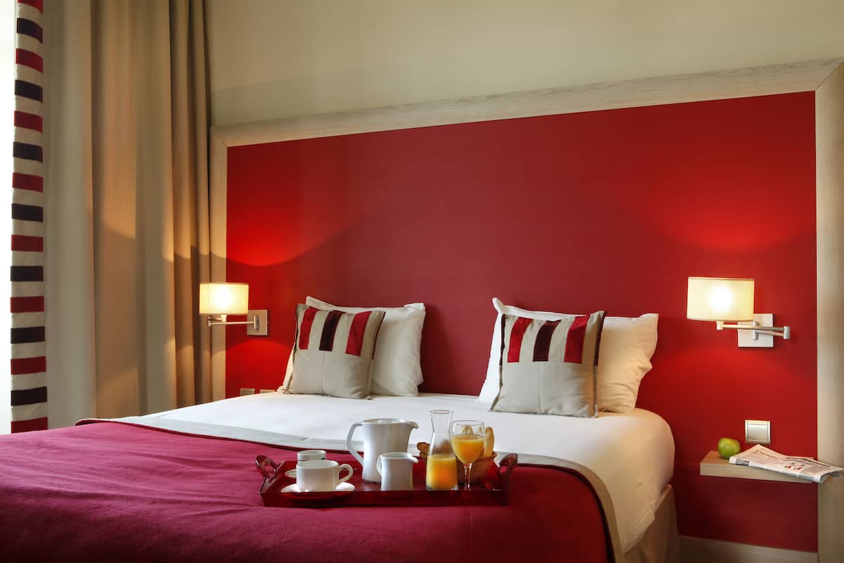 Quarto de hotel com parede de destaque vermelha, cama com roupa de cama branca, duas luminárias de cabeceira, um tabuleiro com itens de café da manhã e uma mesinha de cabeceira com abajur e jornal.
