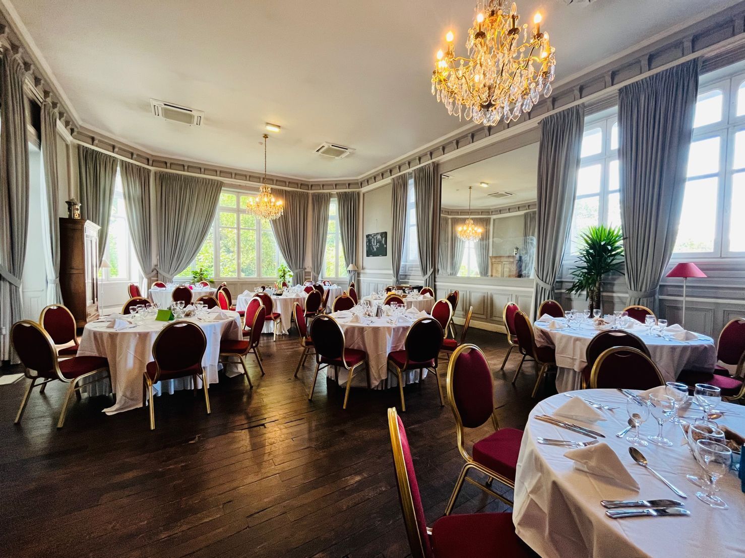 Un comedor elegante con varias mesas redondas dispuestas para una comida, ventanas amplias con cortinas y un candelabro.