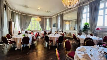 Un comedor elegante con varias mesas redondas dispuestas para una comida, ventanas amplias con cortinas y un candelabro.