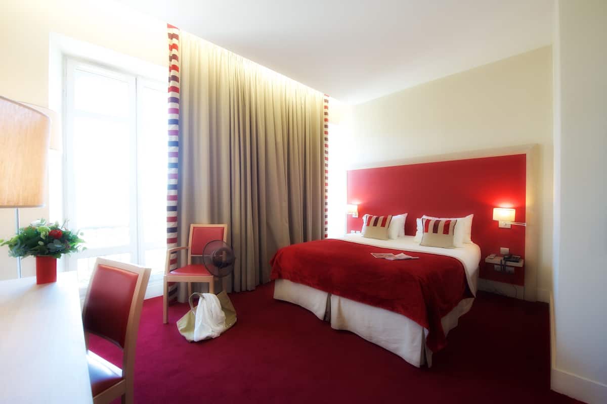 Quarto de hotel com cama, escrivaninha, cadeira, mesa de cabeceira e janela com cortinas.