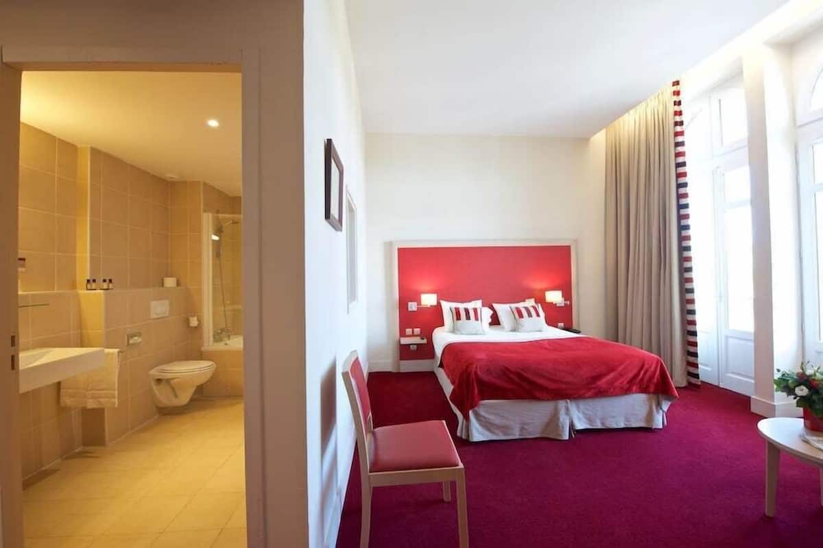 Quarto de hotel com cabeceira vermelha, uma cama de solteiro, mesa de cabeceira, uma cadeira, uma pequena mesa, janela com cortinas e banheiro com chuveiro e vaso sanitário.