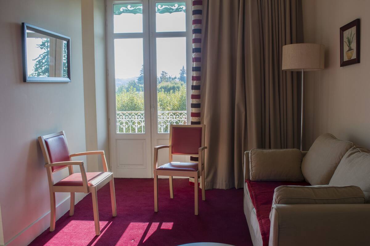 Um quarto com tapete vermelho, duas cadeiras de madeira, um sofá bege, um abajur, uma janela com cortinas e um quadro na parede.