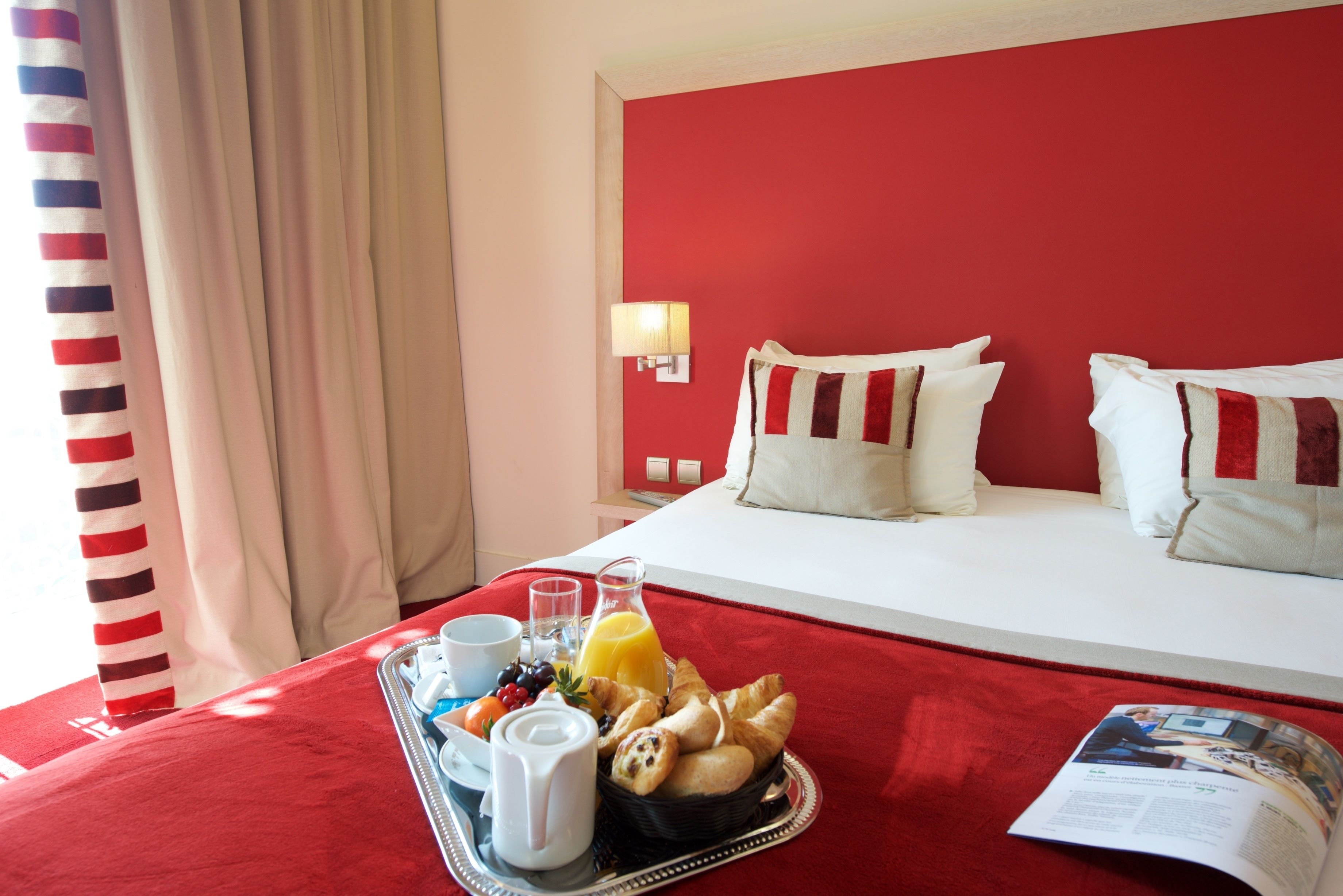 Hotelkamer met een rode wand, een bed met witte lakens, een dienblad met ontbijtartikelen en een tijdschrift op het bed.