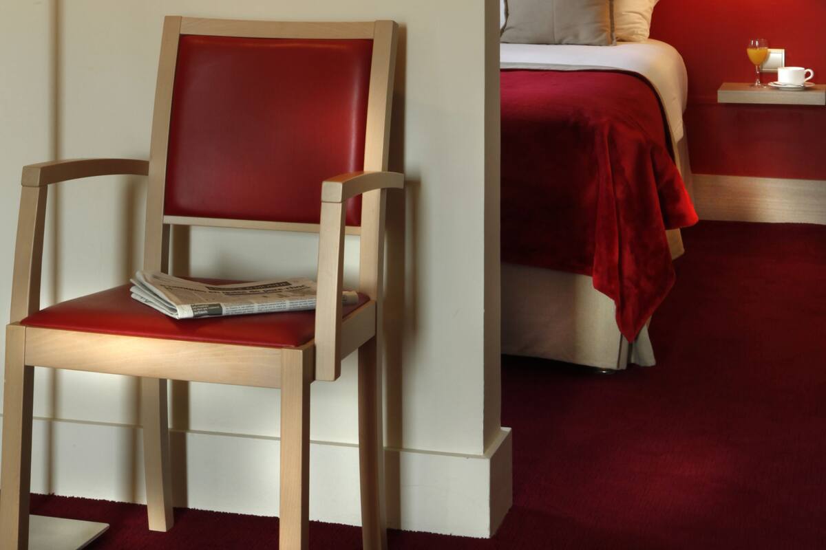 Quarto de hotel com uma cadeira de madeira, uma cama com roupa de cama vermelha, uma mesa de cabeceira com abajur e um quadro pendurado na parede.