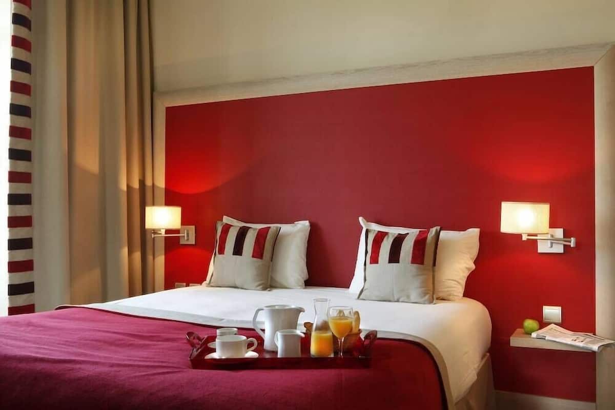 Quarto de hotel com uma parede em vermelho, cama com roupa de cama branca, duas luminárias de cabeceira, bandeja com itens de café da manhã e mesa de cabeceira com abajur.