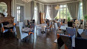 Un comedor amplio con varias mesas y sillas, ventanas grandes con cortinas y un candelabro.